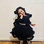toddler, child, smile, black_dress, dress, pose, flooring, wood_floor, wall, happy, cute, girl, standing, footwear, gold_shoes, bracelet, curly_hair, indoors, portrait, fashion