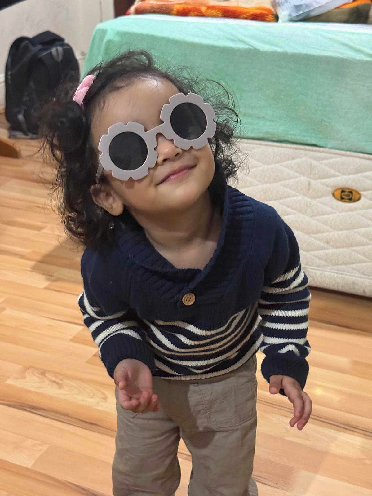 Anand joined the competition — help win amazing prizes! child, toddler, sunglasses, glasses, striped_sweater, curly_hair, indoor, wooden_floor, bed, backpack, smile, playful, cute, clothing, person, young_child, casual, portrait, funny_glasses, home