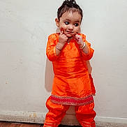 Anand joined the competition — help win amazing prizes! child, orange_clothing, traditional_outfit, standing, dimples, cheeky_pose, wall, floor, wooden_floor, footwear, indoor, cute, smiling, young_child, person, hair_tied, silver_bracelet, candid, full_body, portrait