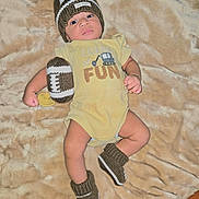 Ezrah joined the competition — help win amazing prizes! baby, infant, knitted_hat, knitted_booties, knitted_football, yellow_onesie, blanket, lying_down, soft_texture, cute, child, indoor, cozy, newborn, toddler, face, expression, warm_clothing, comfort, portrait