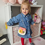 Capucine a rejoint le concours — aidez-le/la à gagner de superbes lots ! toddler, child, denim_dress, red_tights, purse, toy_rabbit, stuffed_toy, bookshelf, children_books, carpet, heart_pattern, indoor, smiling, playful, shoes, floor, toy, person, cute, home