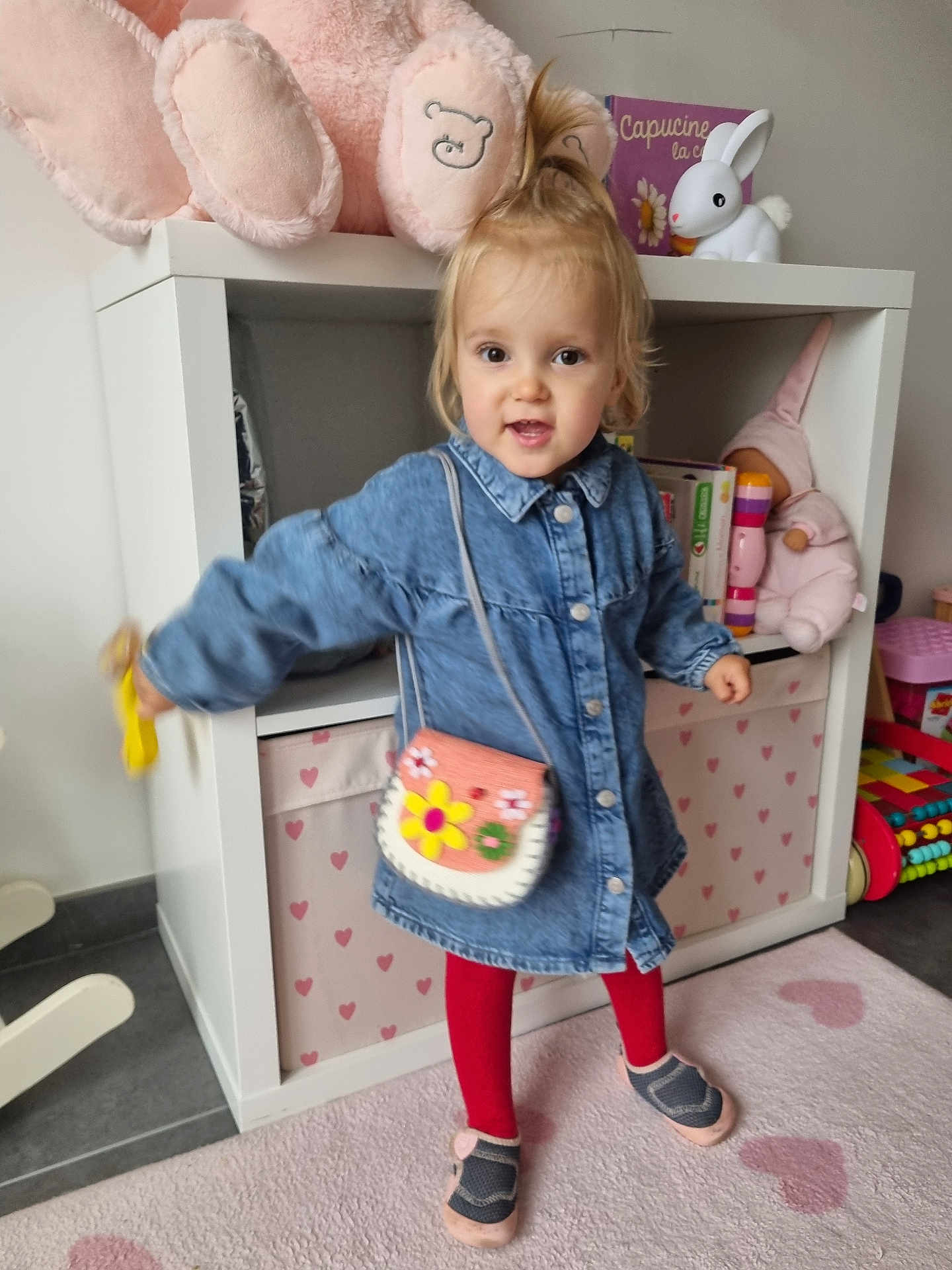 Capucine a rejoint le concours — aidez-le/la à gagner de superbes lots ! toddler, child, denim_dress, red_tights, purse, toy_rabbit, stuffed_toy, bookshelf, children_books, carpet, heart_pattern, indoor, smiling, playful, shoes, floor, toy, person, cute, home