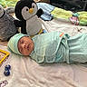 baby, newborn, sleeping, swaddled, plush_toys, penguin_plush, blanket, bed, soft_toys, child, infant, cute, peaceful, indoor, cozy, hat, clothing, resting, nursery, comfort