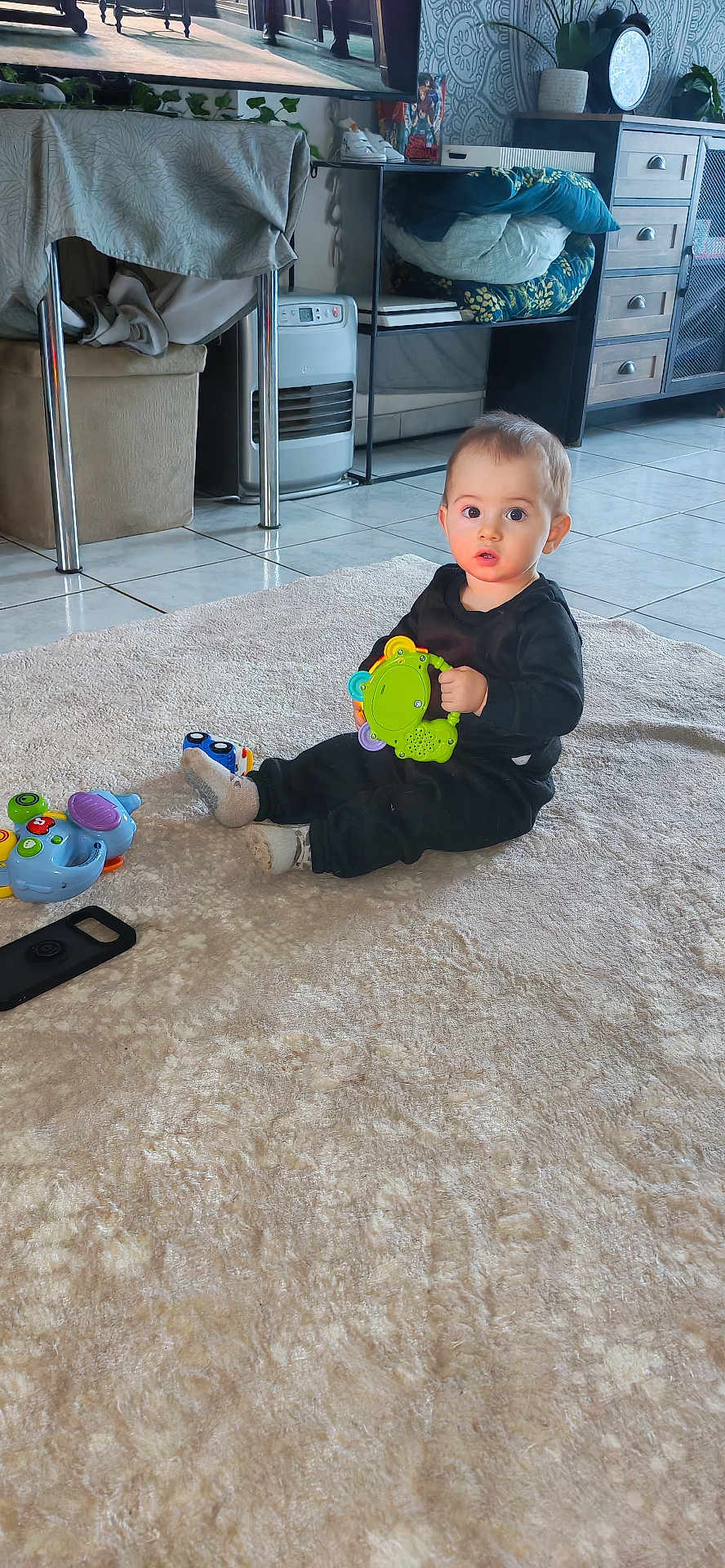 Jérémy a rejoint le concours — aidez-le/la à gagner de superbes lots ! toddler, child, toy, rug, black_clothing, indoor, floor_tiles, furniture, plant, television, cabinet, curious, sitting, playful, colorful_toy, home, living_room, person, young_child, expression