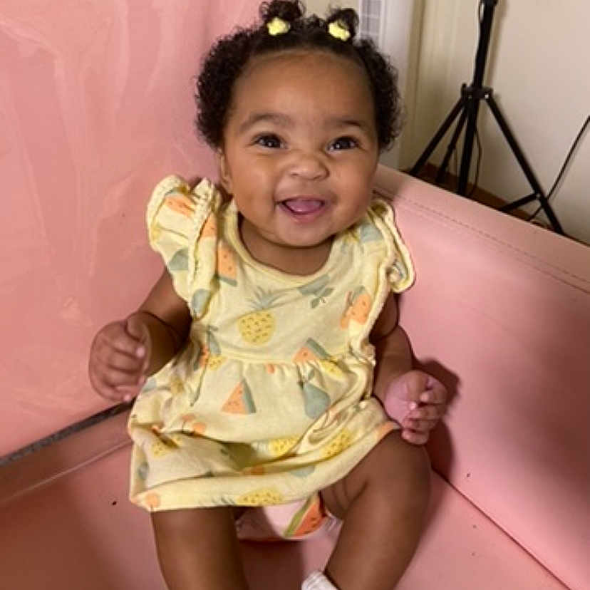 Mylasia joined the competition — help win amazing prizes! baby, child, clothing, cute, expression, face, furniture, hair_buns, happy, indoor, infant, person, pigtails, pink_bench, portrait, seated, smiling, socks, yellow_dress, young_child