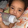 baby, blanket, bottle, child, close_up, curly_hair, expression, face, feeding, floral_pattern, indoors, infant, lying_down, milk, person, pillow, resting, skin, soft_texture, young