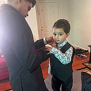 Adam is registered to the contest to win money with this photo: toddler, child, adult, indoor, living_room, shirt, vest, black_pants, socks, curly_hair, door, shoes, carpet, table, bag, person, floor, home, cuff, adjusting_clothes