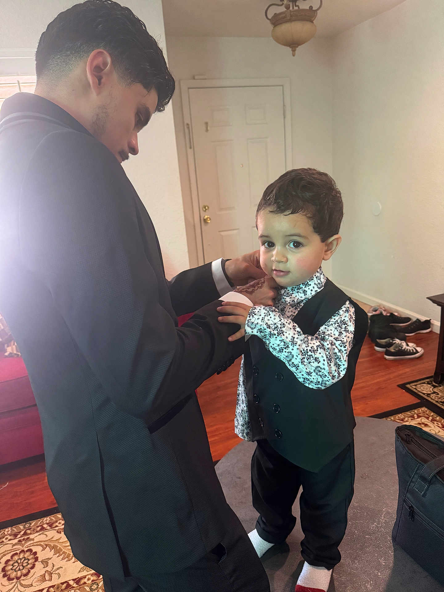 Adam is registered to the contest to win money with this photo: toddler, child, adult, indoor, living_room, shirt, vest, black_pants, socks, curly_hair, door, shoes, carpet, table, bag, person, floor, home, cuff, adjusting_clothes