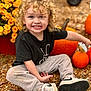 Asher is registered to the contest to win money with this photo: child, toddler, curly_hair, smiling, black_tshirt, gray_pants, white_sneakers, pumpkin, yellow_flowers, outdoor, autumn, gravel, sitting, happy, cute, playful, nature, fall, seasonal, portrait
