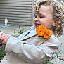 Asher is registered to the contest to win money with this photo: child, toddler, curly_hair, beige_jacket, flower, orange_flower, outdoor, smiling, happy, plant, greenery, concrete, house_siding, side_view, cute, portrait, person, nature, casual, daylight