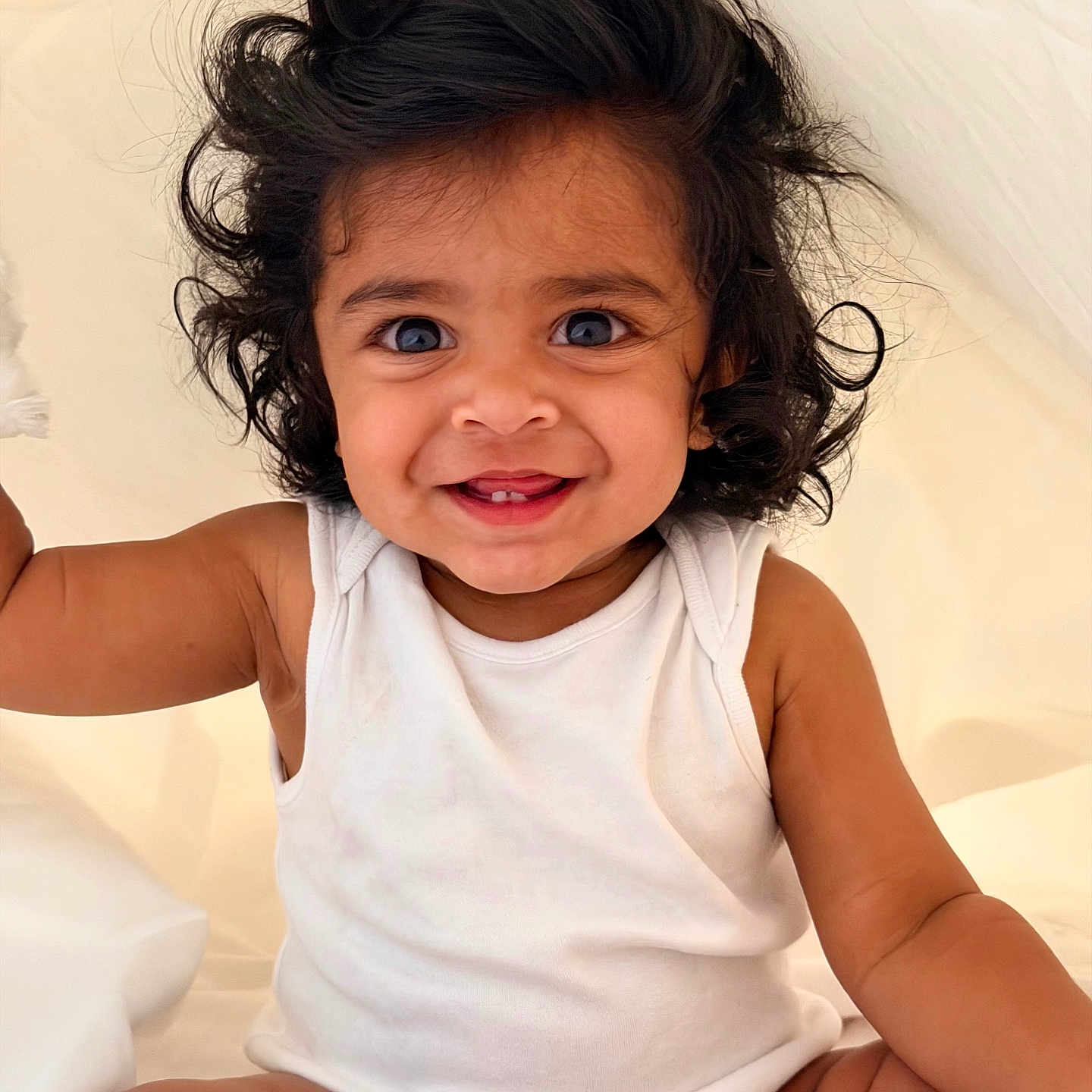 Dheera joined the competition — help win amazing prizes! baby, child, smiling, curly_hair, white_onesie, sitting, portrait, cute, happy, infant, face, skin, arm, indoors, light_background, baby_toddler, person, young_child, expression, closeup