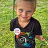 child, boy, birthday, outdoor, grass, smiling, face, blue_eyes, black_tshirt, colorful_print, pants, shoes, dirt, playful, celebration, badge, standing, casual_clothing, daylight, fun