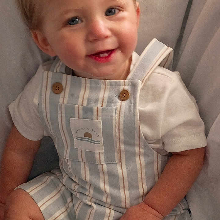 Noah is registered to the contest to win money with this photo: arm, baby, background, blonde_hair, child, clothing, cute, face, happy, indoor, leg, overalls, person, portrait, sitting, skin, smiling, toddler, white_shirt, young_child
