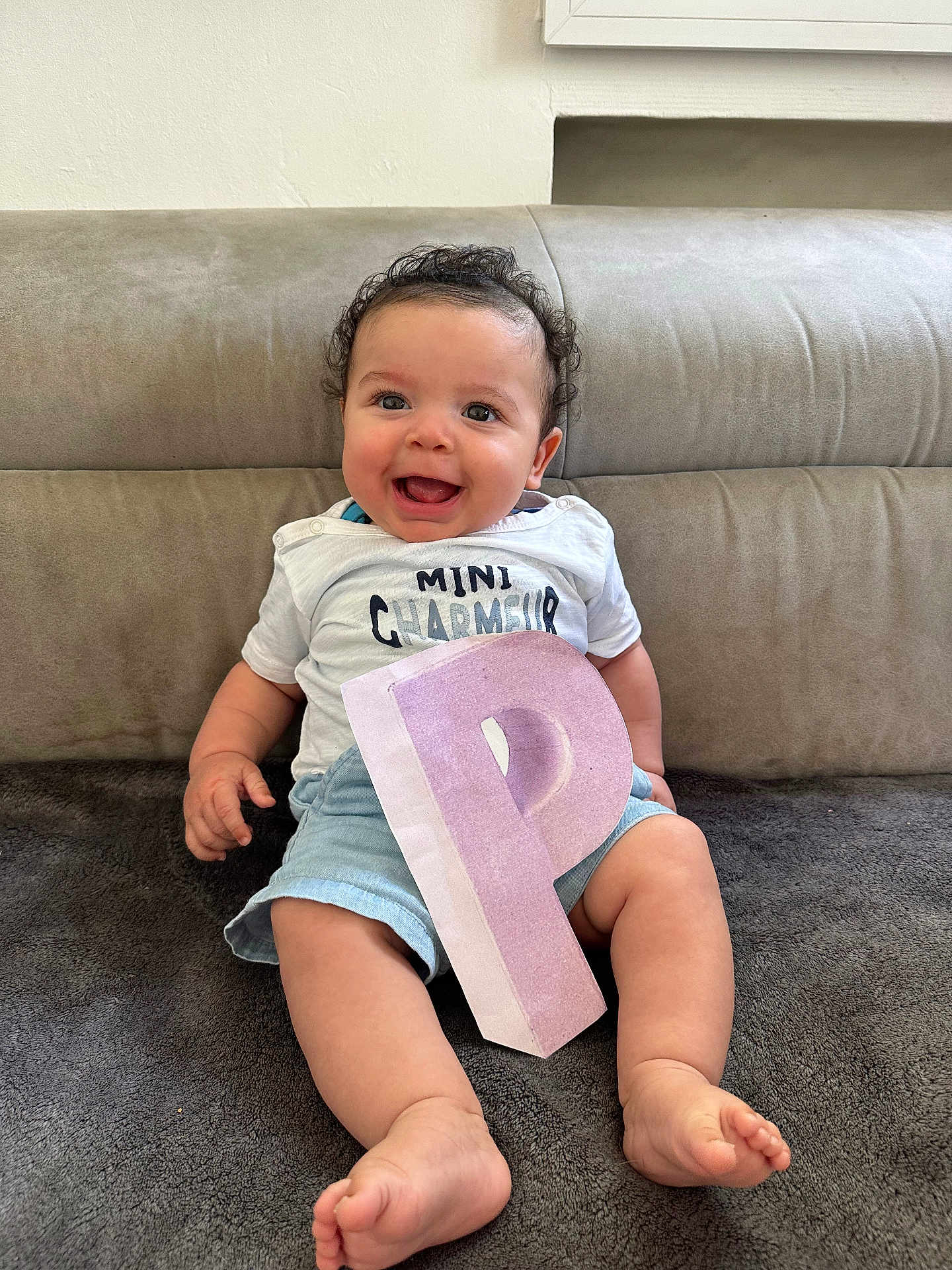 Kayden a rejoint le concours — aidez-le/la à gagner de superbes lots ! baby, child, smiling, curly_hair, white_shirt, blue_shorts, letter_p, pink_letter, sitting, blanket, couch, happy, indoors, portrait, cute, feet, hand, face, person, young_child