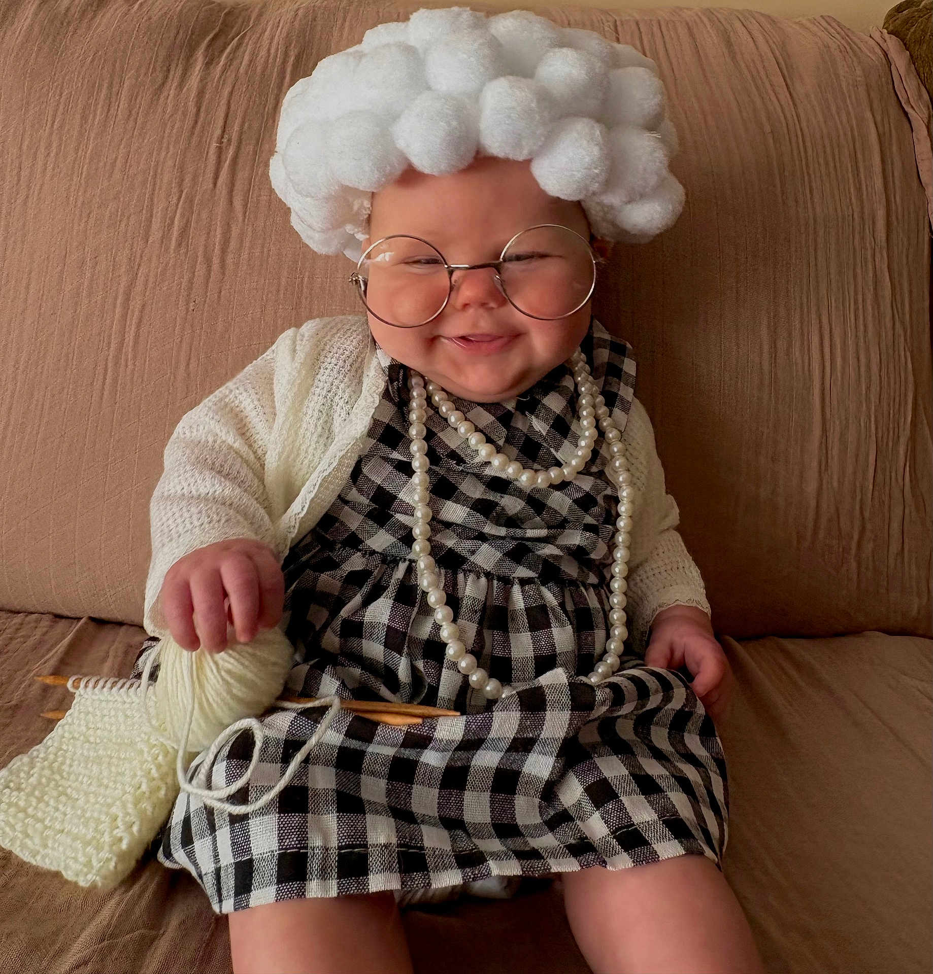 Emory joined the competition — help win amazing prizes! baby, child, costume, glasses, knitting, yarn, wool, pearls, plaid_dress, couch, smile, indoors, cute, funny, handmade, white_hair_wig, knitting_needles, cozy, sitting, portrait