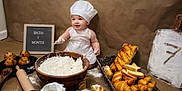 Enzo joined the competition — help win amazing prizes! baby, child, chef_hat, apron, flour, eggs, bread, croissants, muffins, rolling_pin, wooden_spoon, basket, baking, kitchen, smile, sitting, table, brown_background, sign, portrait