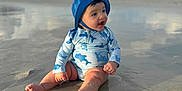 Enzo is registered to the contest to win money with this photo: baby, beach, blue, child, clouds, daytime, happy, hat, nature, outdoor, person, reflection, sand, shore, sitting, smiling, sunny, swimsuit, vacation, water