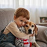 child, dog, hug, couch, indoor, denim_overalls, stuffed_toy, curtains, soft_light, portrait, pet, cozy, home, comfort, friendship, casual_clothing, brown_hair, animal, toddler, affection