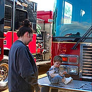 Johnathan is registered to the contest to win money with this photo: child, woman, fire_truck, vehicle, outdoor, daylight, person, face_paint, sneakers, jeans, jacket, reflection, metal, bumper, equipment, smile, side_view, casual_clothing, curly_hair, interaction