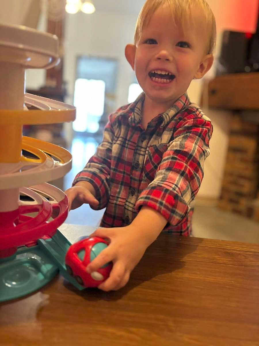 Tiago participe au concours pour gagner de l'argent avec cette photo : toddler, child, smiling, plaid_shirt, toy_car, wooden_table, indoor, playing, happy, face, hands, person, furniture, bright, light, background, blur, young_child, cute, casual