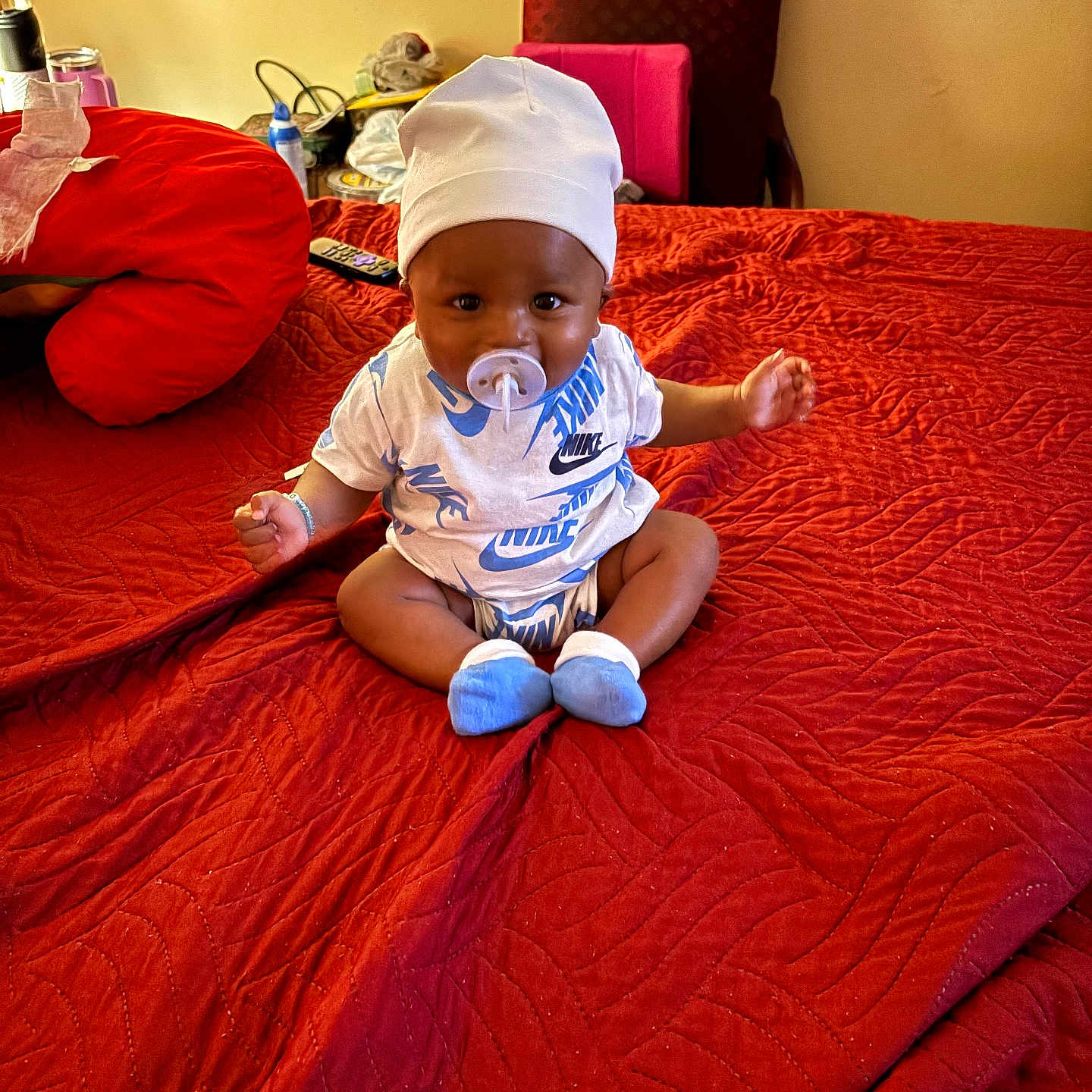 Demitrius Jr. is registered to the contest to win money with this photo: baby, blanket, bottle, chair, child, clothing, cute, footwear, home, indoor, infant, nike_shirt, pacifier, person, red_bedspread, remote_control, sitting, toy, wall, white_cap