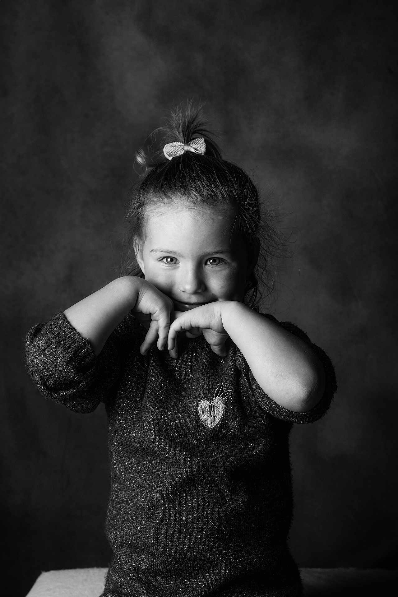 Julia participe au concours pour gagner de l'argent avec cette photo : child, girl, portrait, black_and_white, studio, smile, bow, sweater, hands, eyes, hair, face, young_child, indoor, playful, pose, seated, backdrop, cute, expression