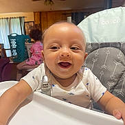 Tiberias is registered to the contest to win money with this photo: baby, child, high_chair, smiling, indoor, onesie, dinosaur_pattern, person, furniture, curtains, ceiling_fan, wood_paneling, laundry_basket, pajamas, happy, infant, seat, room, home, toddler