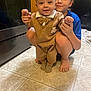 baby, child, older_child, kitchen, floor, tile_floor, brown_outfit, bow_tie, socks, barefoot, smiling, holding_hands, supporting, indoor, person, cute, happy, family, helping, crouching