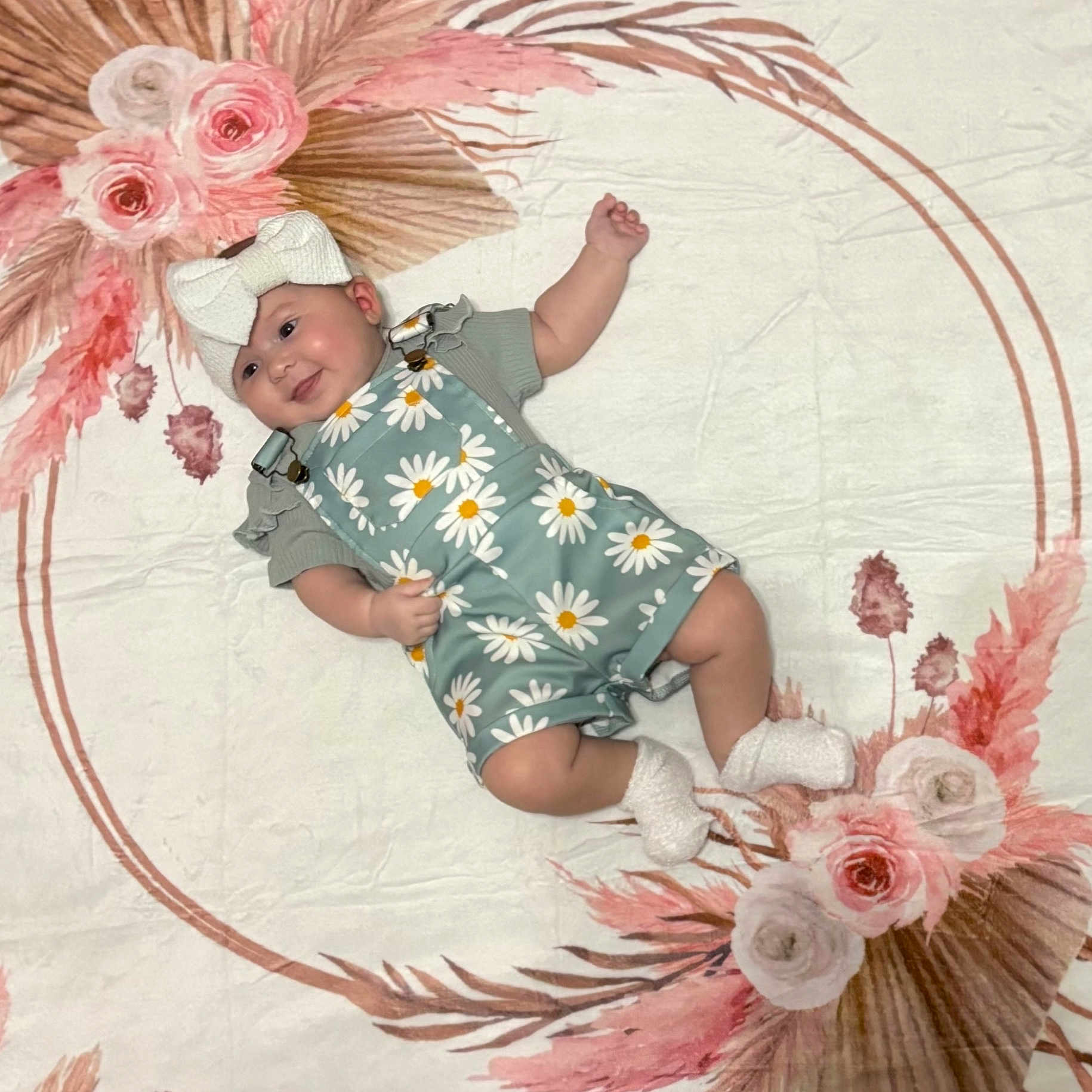 Shetland is registered to the contest to win money with this photo: baby, blanket, bow, child, cute, daisy_print, floral_pattern, green, happy, headband, indoors, infant, lying_down, overalls, pink, portrait, smile, socks, soft_texture, white