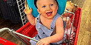 Phoenix is registered to the contest to win money with this photo: toddler, shopping_cart, hat, child, smiling, blue_hat, overalls, store, clothing, aisle, floor, happy, baby, cute, playing, retail, colorful, metal, footwear, girl