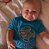 baby, child, smiling, blue_shirt, shirt_print, bed, bedding, blanket, person, infant, cute, happy, lying_down, human, skin, shorts, indoor, young_child, face, arm