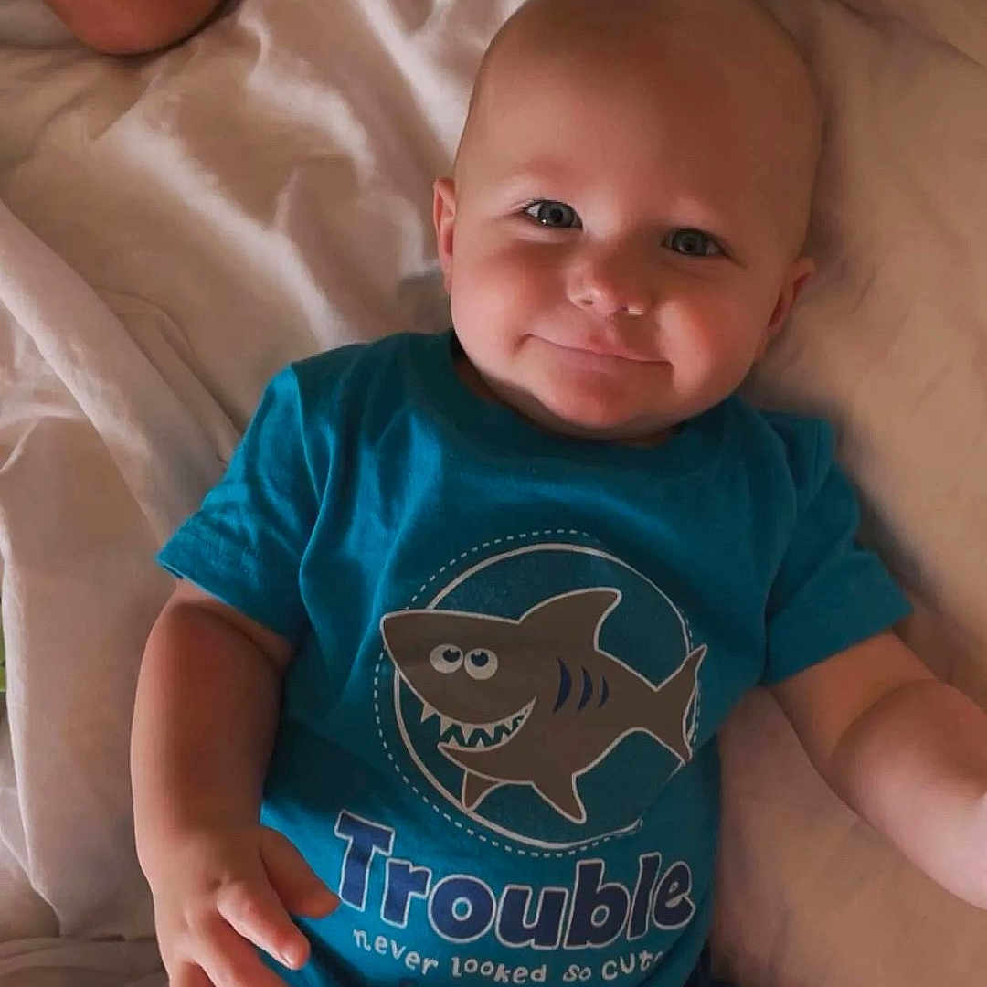 Matthew joined the competition — help win amazing prizes! baby, child, smiling, blue_shirt, shirt_print, bed, bedding, blanket, person, infant, cute, happy, lying_down, human, skin, shorts, indoor, young_child, face, arm