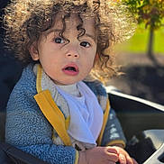 Brooks joined the competition — help win amazing prizes! toddler, child, curly_hair, portrait, face, surprised, open_mouth, eyes, sunlight, outdoor, car, car_seat, jacket, yellow_clothing, blue_fleece, bib, hands, steering_wheel, greenery, close_up