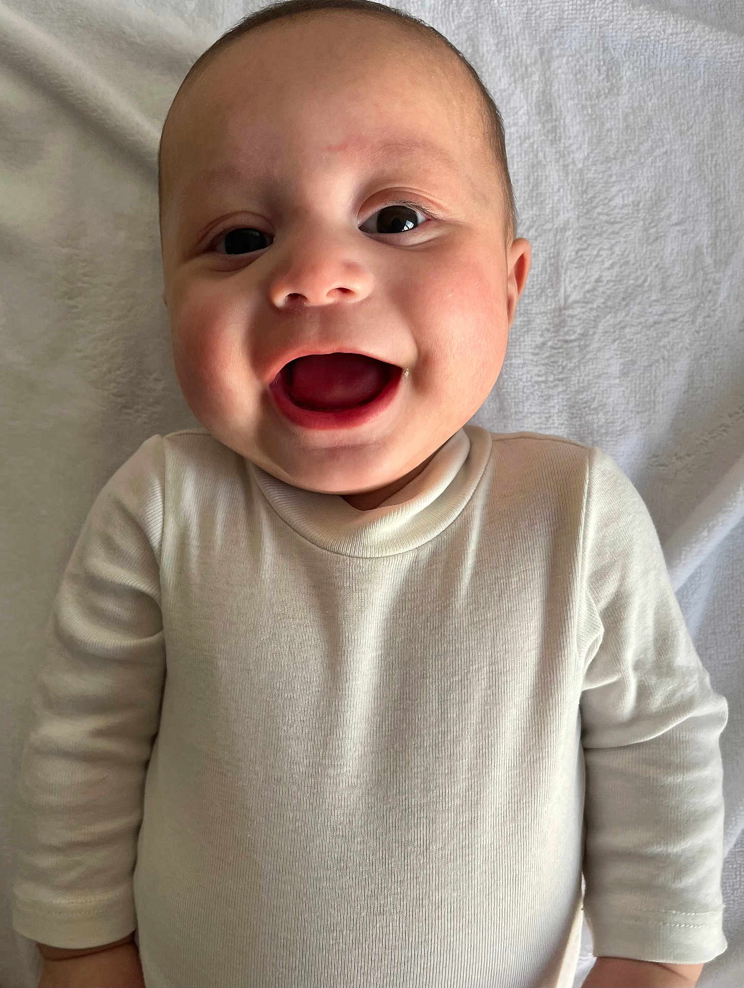 Taimanarii participe au concours pour gagner de l'argent avec cette photo : baby, infant, smile, open_mouth, onesie, white_clothing, face, cheeks, big_eyes, portrait, close_up, blanket, towel, lying_down, happy, cute, skin, head, ear, hands