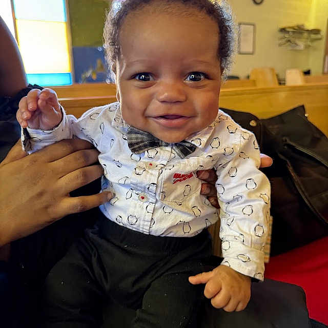 Dymier joined the competition — help win amazing prizes! baby, child, smiling, indoor, colorful, stained_glass, shirt, bow_tie, person, happy, cute, portrait, hands, seated, black_pants, cozy, warm_lighting, infant, clothing, background