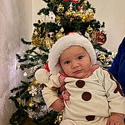 Nëlyo a rejoint le concours — aidez-le/la à gagner de superbes lots ! baby, child, christmas_tree, holiday, festive, santa_hat, snowman_outfit, decorations, ornaments, tinsel, indoor, person, cute, face, celebration, seasonal, festivity, holding, christmas, winter