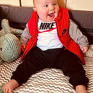 Oliver is registered to the contest to win money with this photo: baby, child, smiling, red_vest, nike_logo, stuffed_animal, indoor, happy, sitting, patterned_blanket, pants, foot, face, clothing, cute, infant, toy, cheerful, portrait, casual