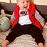 baby, child, smiling, red_vest, nike_logo, stuffed_animal, indoor, happy, sitting, patterned_blanket, pants, foot, face, clothing, cute, infant, toy, cheerful, portrait, casual