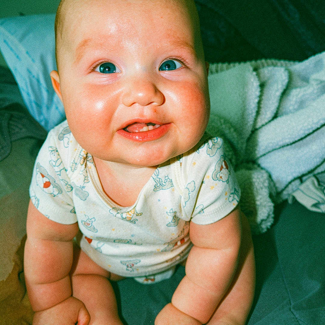 Oliver is registered to the contest to win money with this photo: baby, infant, smiling, blue_eyes, chubby_cheeks, onesie, sitting, blanket, bed, cute, child, portrait, happy, indoors, skin, face, hands, feet, expression, young
