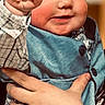 baby, child, infant, face, hand, fist, clothing, blue_outfit, plaid_shirt, holding_hand, person, portrait, cute, smile, indoor, warm_lighting, skin, cheeks, eyes, babywearing