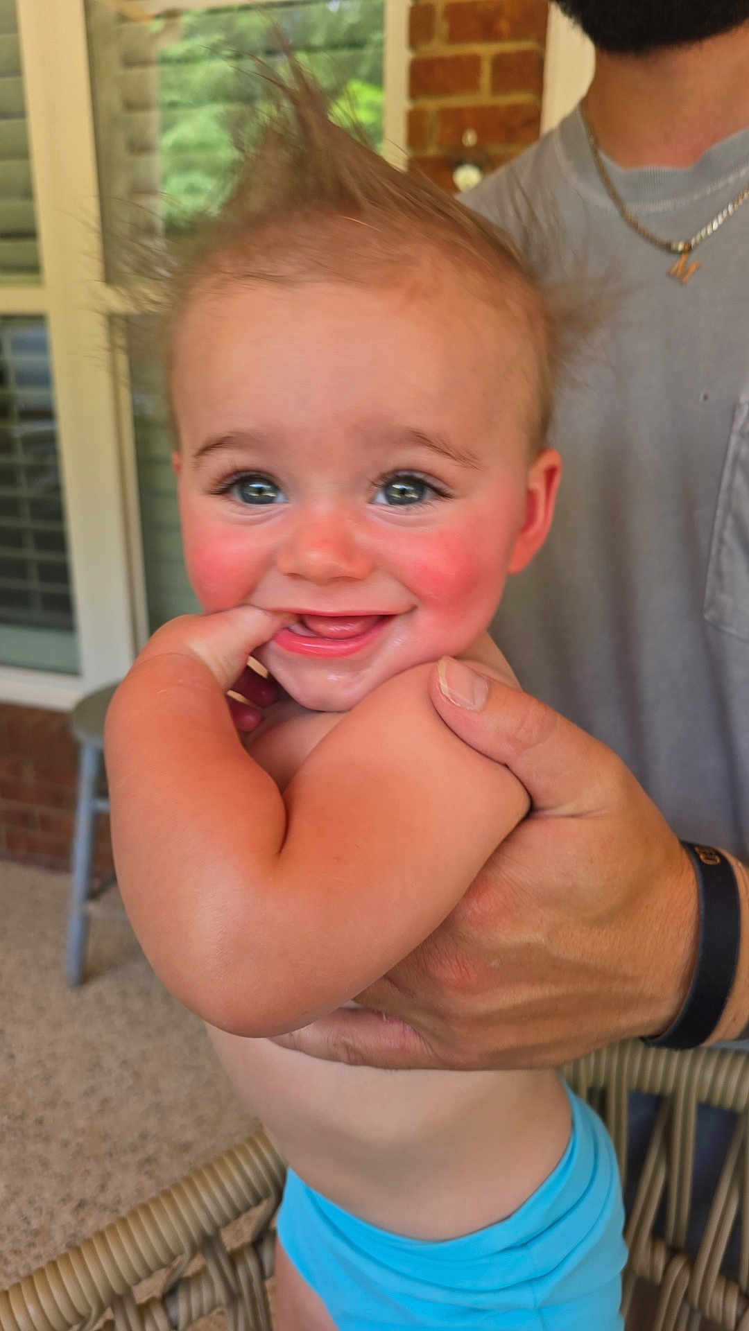 Monty is registered to the contest to win money with this photo: baby, child, smiling, hair, finger, hand, adult_hand, blue_clothing, porch, window, brick_wall, chair, happy, skin, person, closeup, cute, expression, outdoor, holding