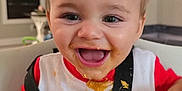 Monty joined the competition — help win amazing prizes! baby, child, high_chair, food, messy, smile, happy, face, bib, indoor, table, cheese, green_beans, person, clothing, red_shirt, white_bib, hair, mouth_open, joy