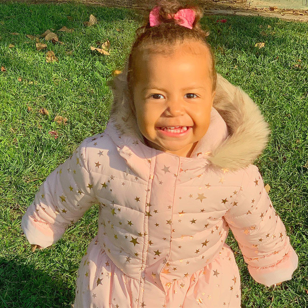 Symphony is registered to the contest to win money with this photo: cheek, child, emotion, eye, face, facial_expression, girl, grass, hair, happiness, head, infant, joy, nose, outerwear, person, pink, skin, smile, standing