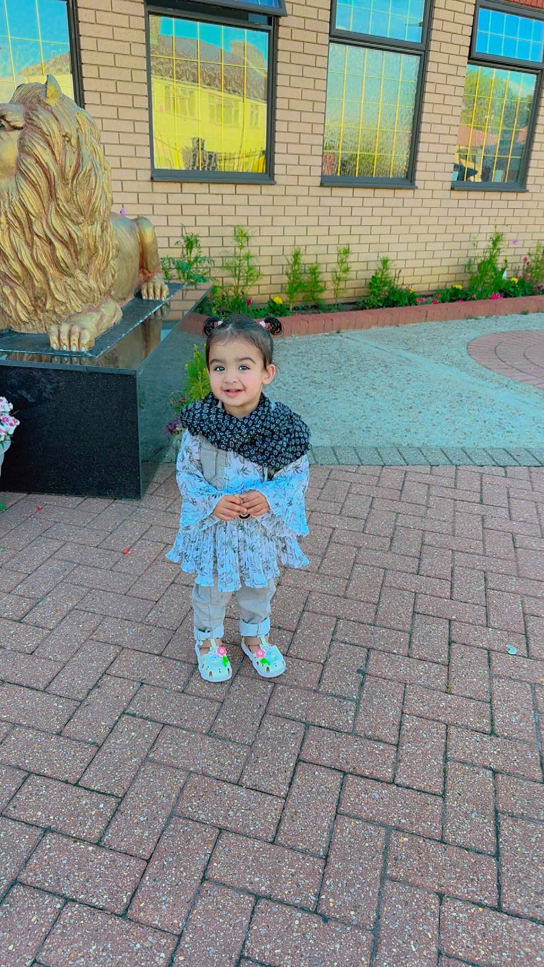 Arveen is registered to the contest to win money with this photo: child, toddler, smiling, outdoor, brick_pavement, statue, lion_statue, building, window, reflection, flower, scarf, clothing, shoes, person, cute, happy, daylight, nature, plant