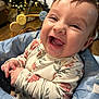 baby, baby_seat, blue_cushion, child, christmas_tree, closeup, cozy, cute, face, floral_clothing, happy, holiday_lights, indoor, infant, laughing, person, smiling, toy, warm_lighting, wooden_floor