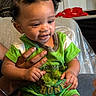 baby, child, green_clothing, hand, couch, indoor, smiling, person, hair_buns, seated, infant, cute, happy, skin, face, holding, text_on_clothing, adult_hand, fabric, home