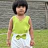 bow, casual, child, daylight, expression, fashion, fence, footwear, grass, green_top, outdoor, person, pink_shoes, portrait, standing, stone_wall, summer_clothing, urban, white_shorts, young_child