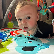 Manuelito is registered to the contest to win money with this photo: baby, infant, play_mat, toys, colorful, curious, face, child, indoors, close_up, soft_toys, black_clothing, play_gym, baby_toys, early_learning, candid, bright_colors, person, cute, exploration