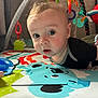 baby, infant, play_mat, toys, colorful, curious, face, child, indoors, close_up, soft_toys, black_clothing, play_gym, baby_toys, early_learning, candid, bright_colors, person, cute, exploration