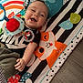 Manuelito is registered to the contest to win money with this photo: baby, infant, smiling, play_mat, toy, colorful, cute, happy, child, indoor, floor, clothing, black_shirt, orange, blue, green, white, pattern, person, reaching